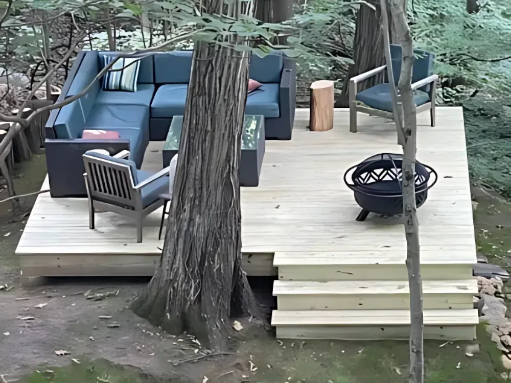Wooden staircase construction leading to a cozy outdoor seating area, featuring a wooden deck with lounge furniture, surrounded by trees.
